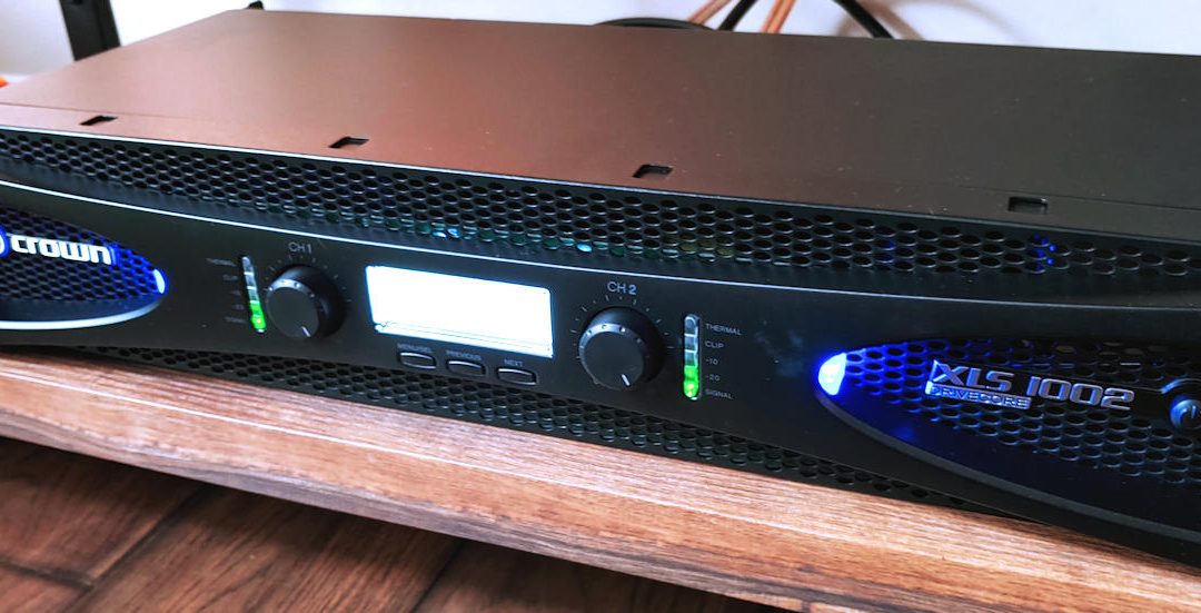 Crown XLS series power amplifier with illuminated front panel controls on a wooden media shelf.